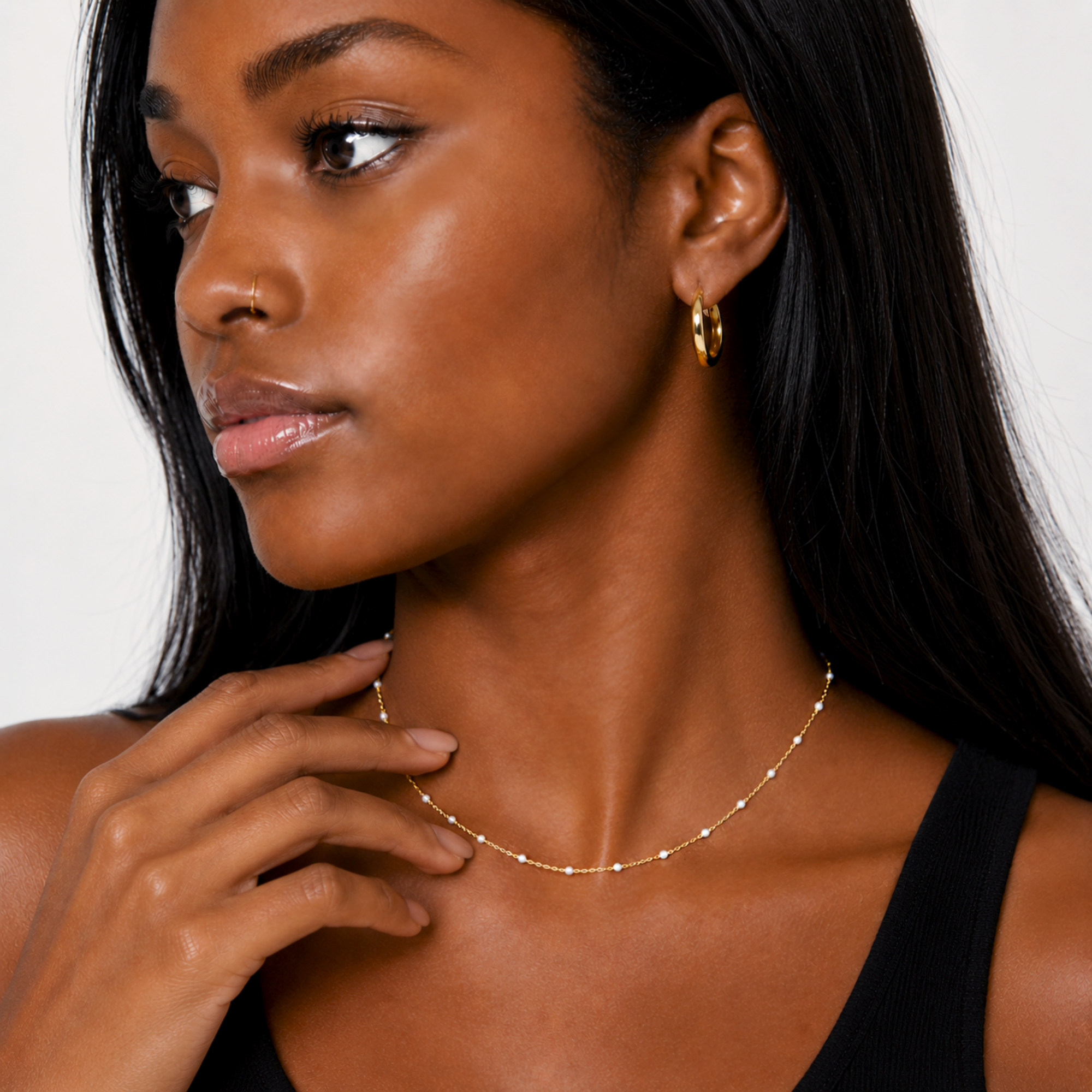 Woman wearing a dainty white beaded gold chain necklace and hoop earrings against a neutral background
