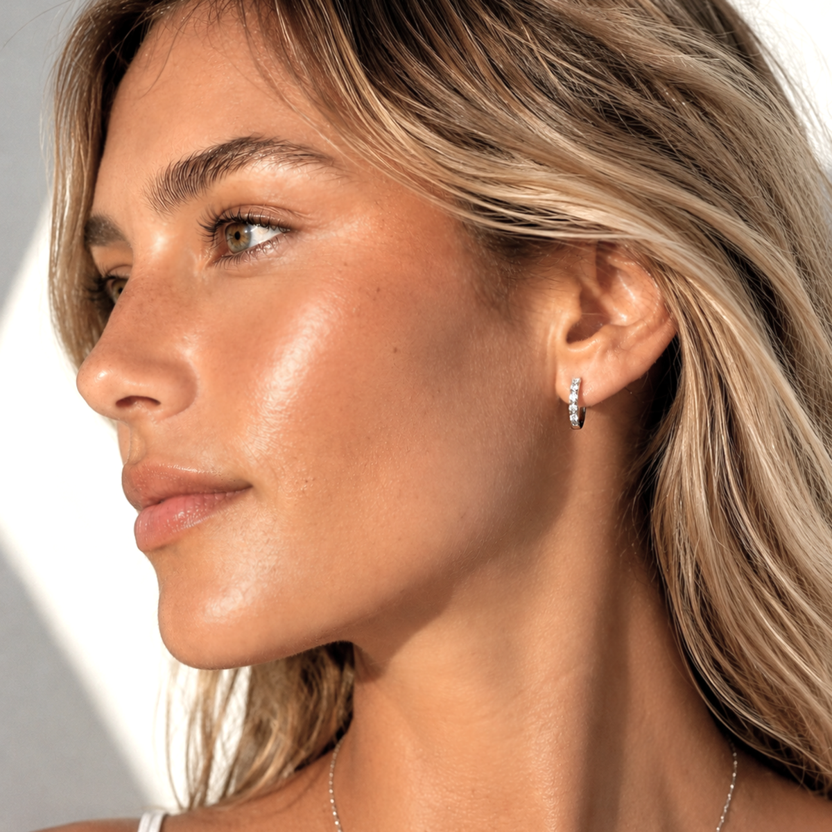 A close-up side profile of a woman with blonde hair wearing a small silver huggie hoop earring with clear stones, set against a bright neutral background.