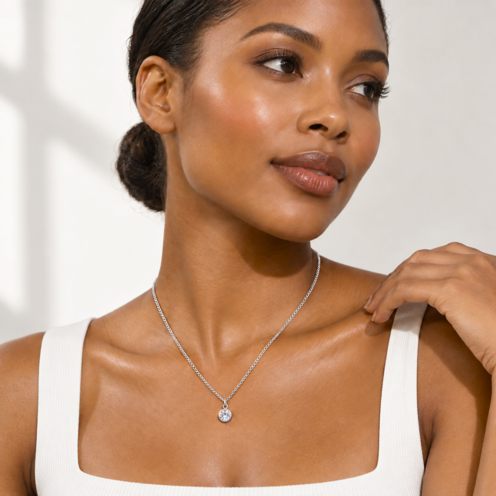 A woman wearing a delicate silver chain necklace with a small round clear gemstone pendant, styled with a white ribbed tank top against a bright, softly lit background.