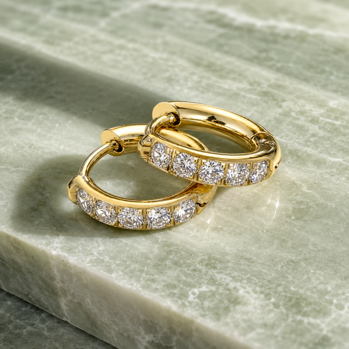 A pair of gold huggie hoop earrings set with round-cut diamonds, resting on a polished green marble surface.