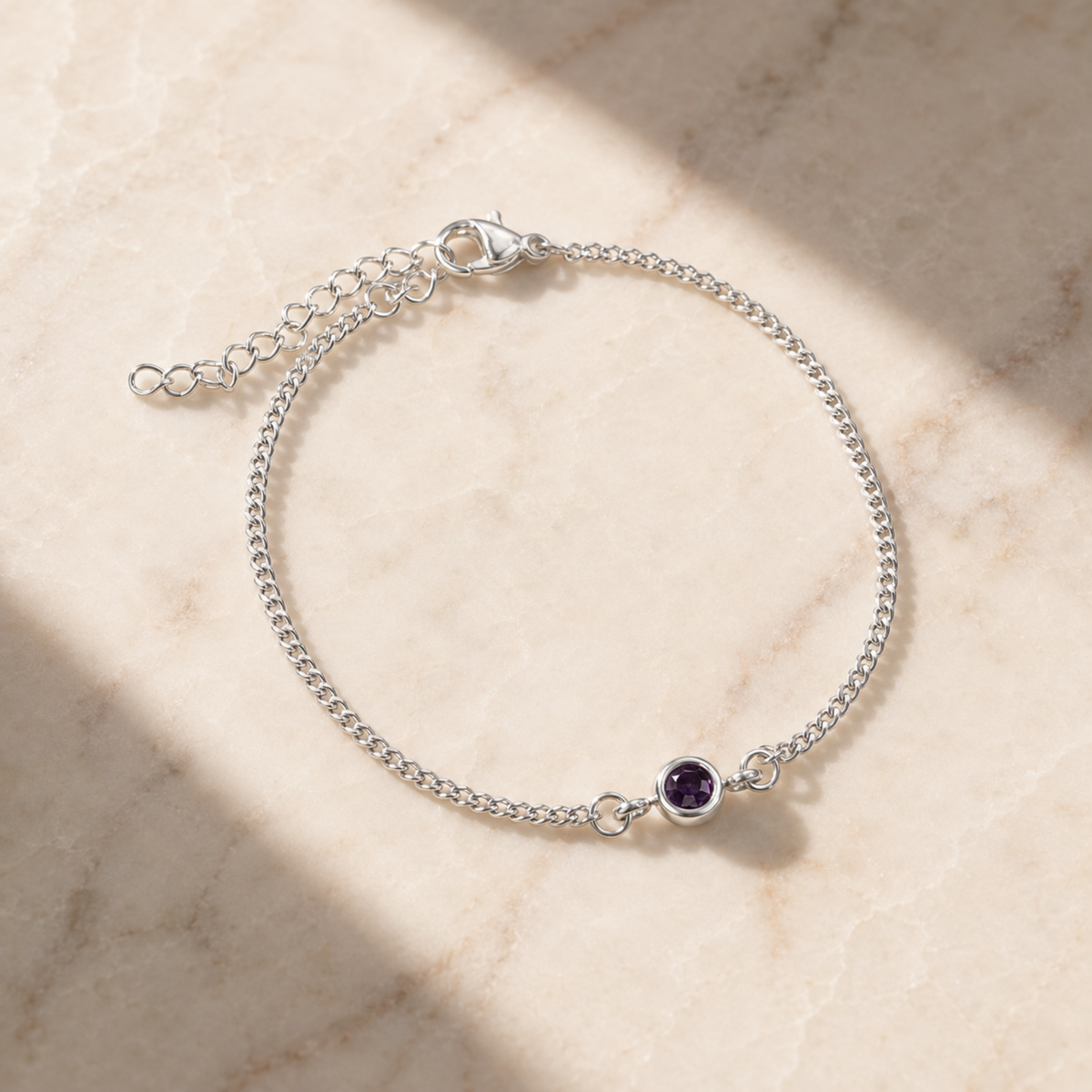 A delicate silver chain bracelet with a small round purple gemstone, displayed on a light beige marble surface with soft shadows.