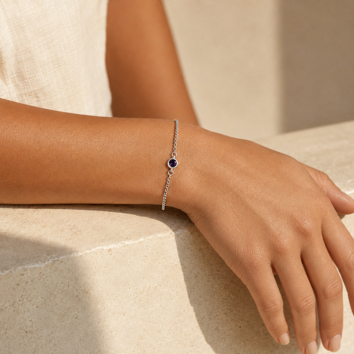 A delicate silver chain bracelet with a central dark purple february birthstone, worn on a woman's wrist resting on a light beige stone surface.