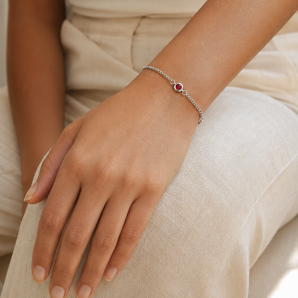 A delicate silver chain bracelet with a small red heart-shaped stone, worn on a person's wrist resting on light beige linen fabric.