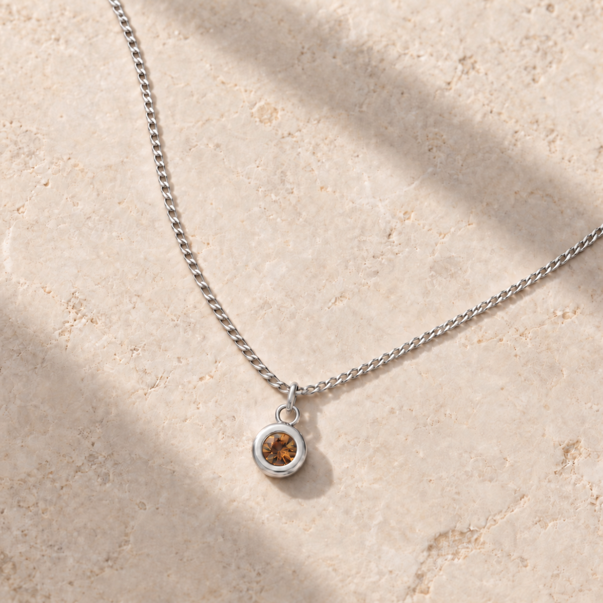 A delicate silver chain necklace with a small round bezel pendant containing a warm amber-brown citrine gemstone, displayed on a textured beige stone surface with soft diagonal shadows. Silver birthstone necklace