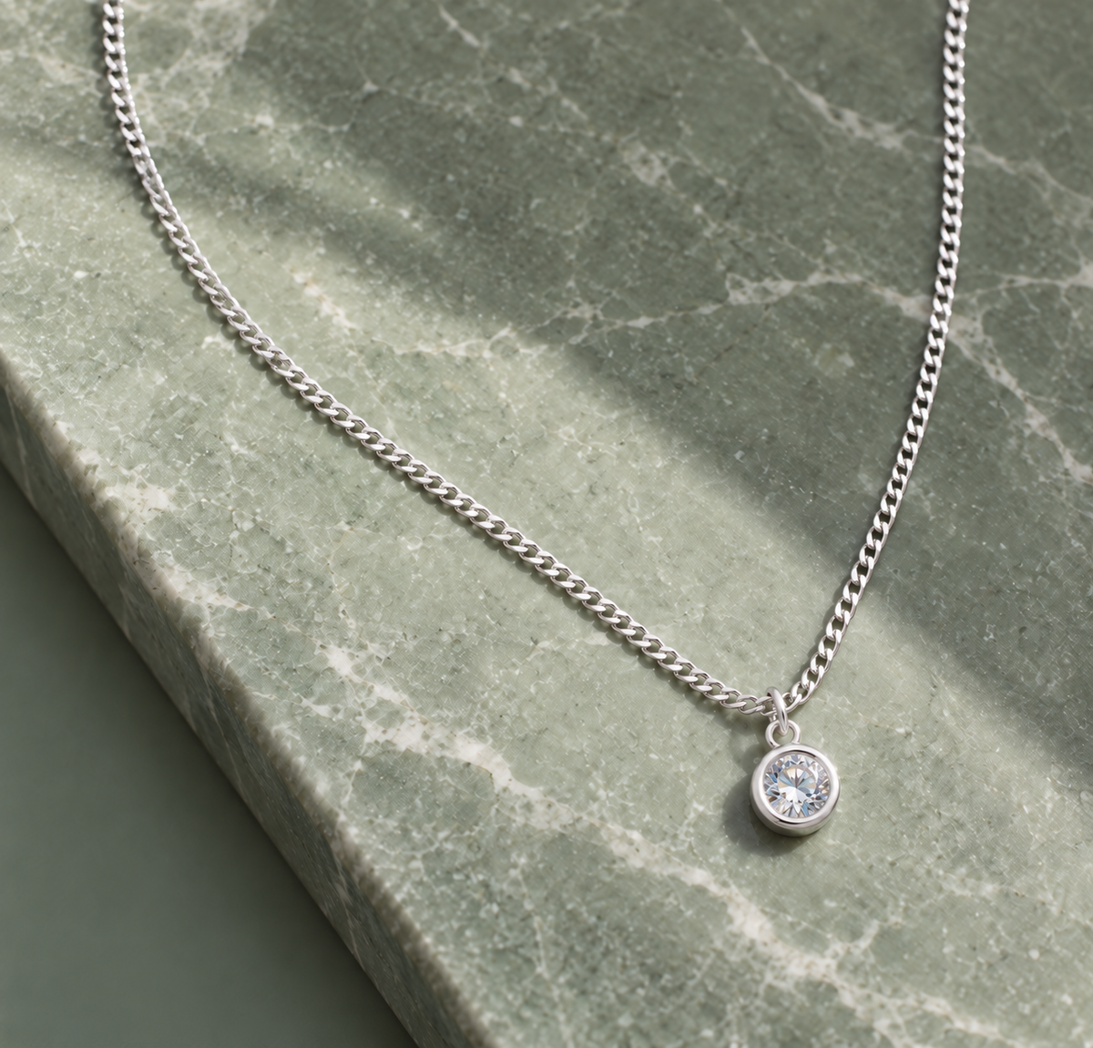 A silver chain necklace with a small round bezel-set clear gemstone pendant, displayed on a green marble surface.