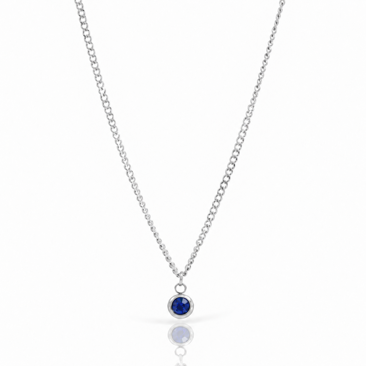 A silver chain necklace with a small round bezel-set deep blue gemstone pendant, displayed on a clean white background with a soft reflection below.