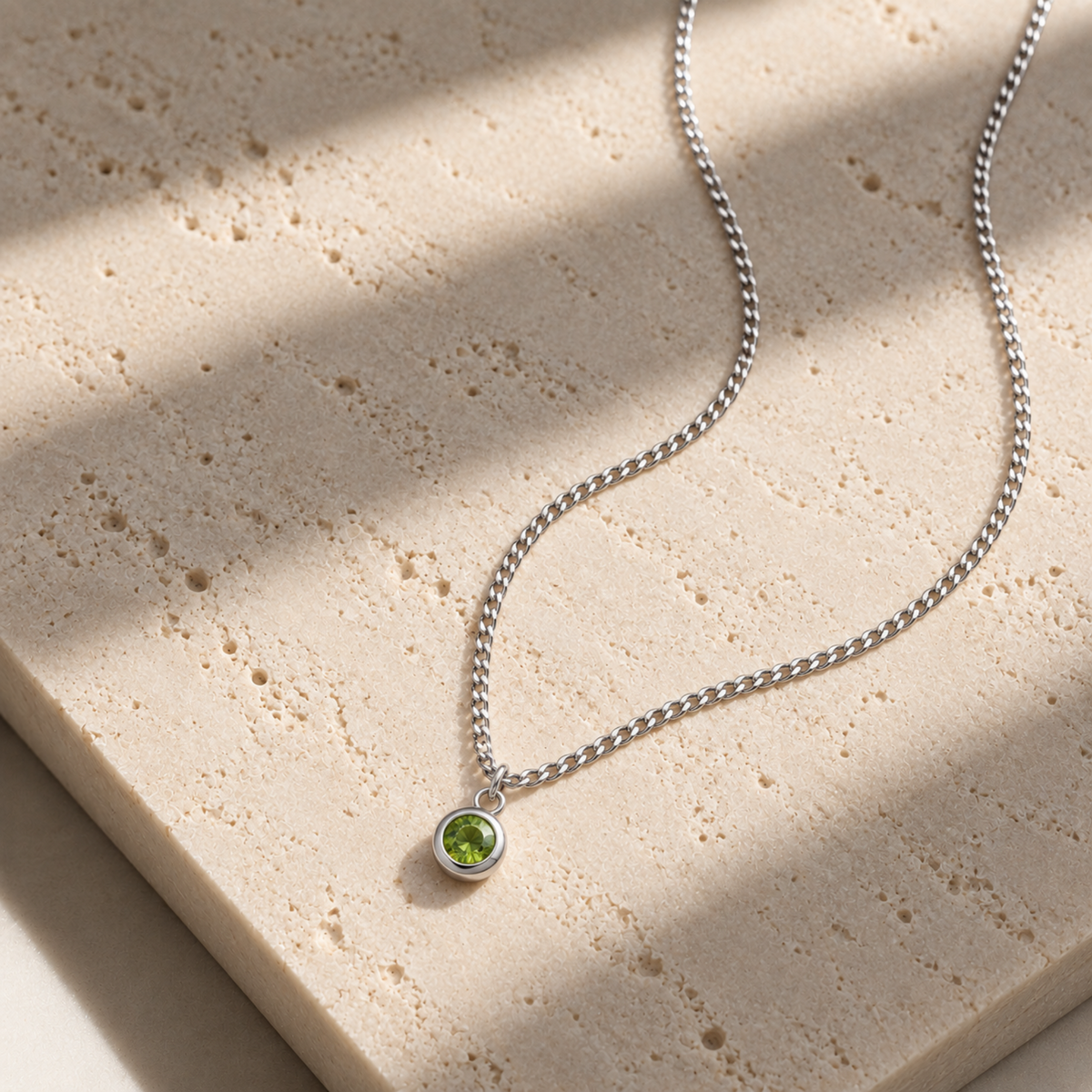 A silver chain necklace with a small round bezel pendant containing a vibrant green gemstone, displayed on a textured beige stone slab with soft diagonal shadows.