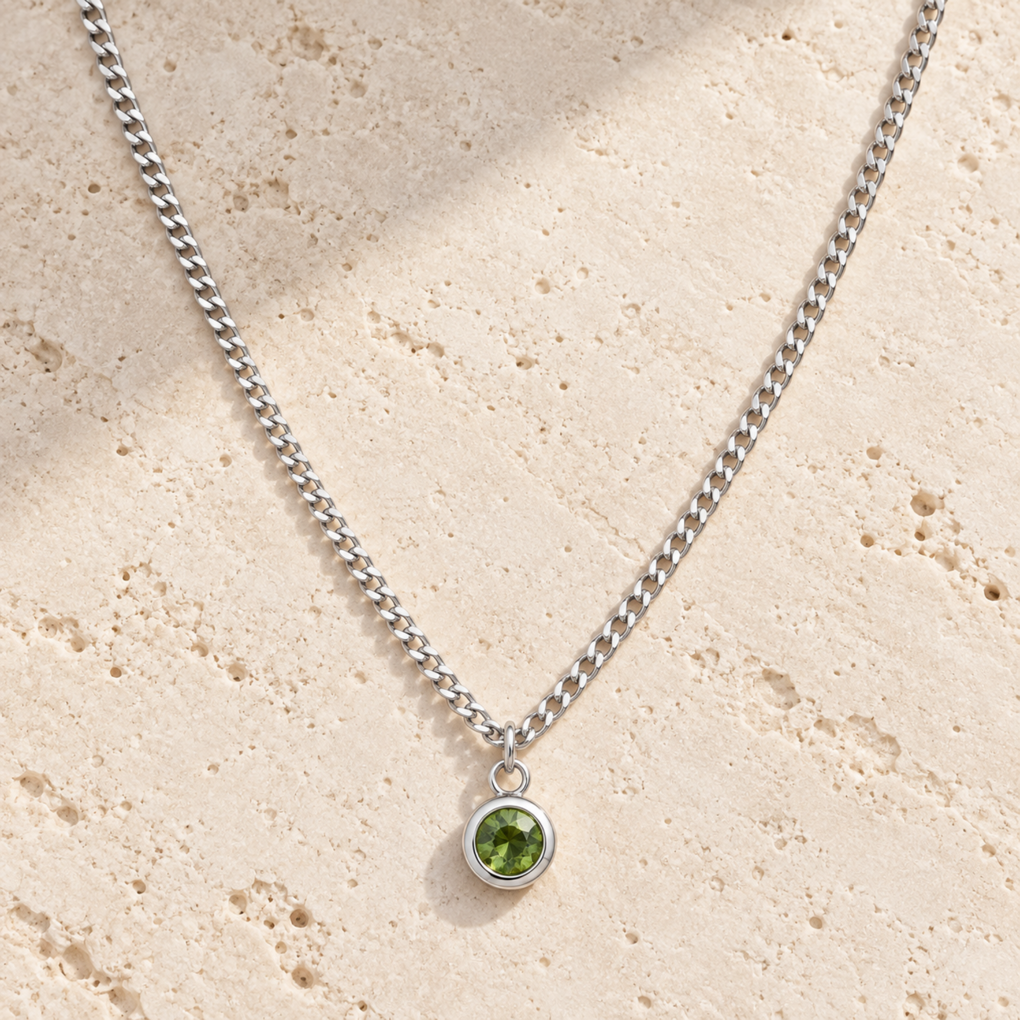 A silver curb chain necklace with a small round bezel pendant featuring a green gemstone, displayed on a textured beige stone surface.