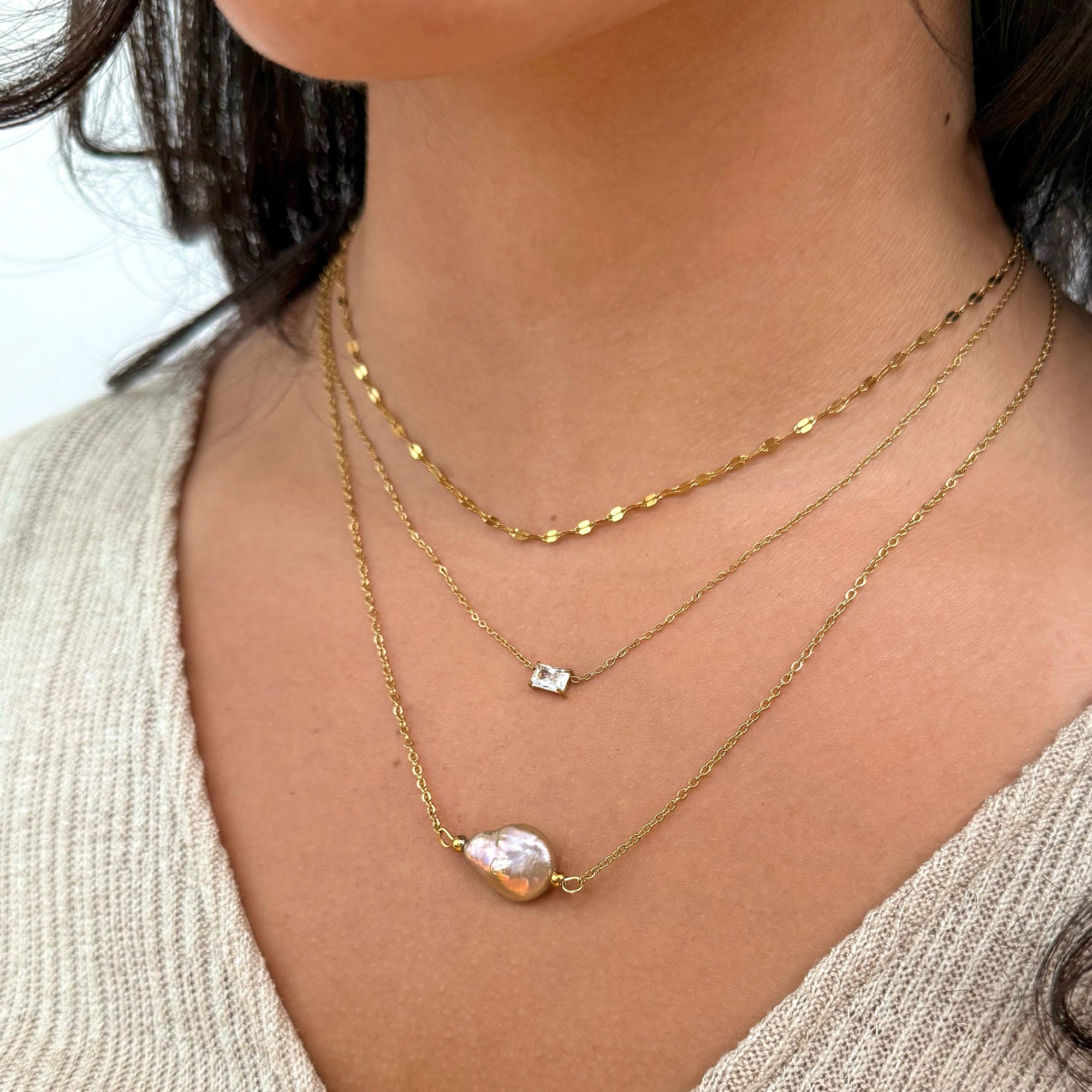 Close-up of a person wearing three gold necklaces with a pearl pendant and a small square pendant and a dainty gold lace chain