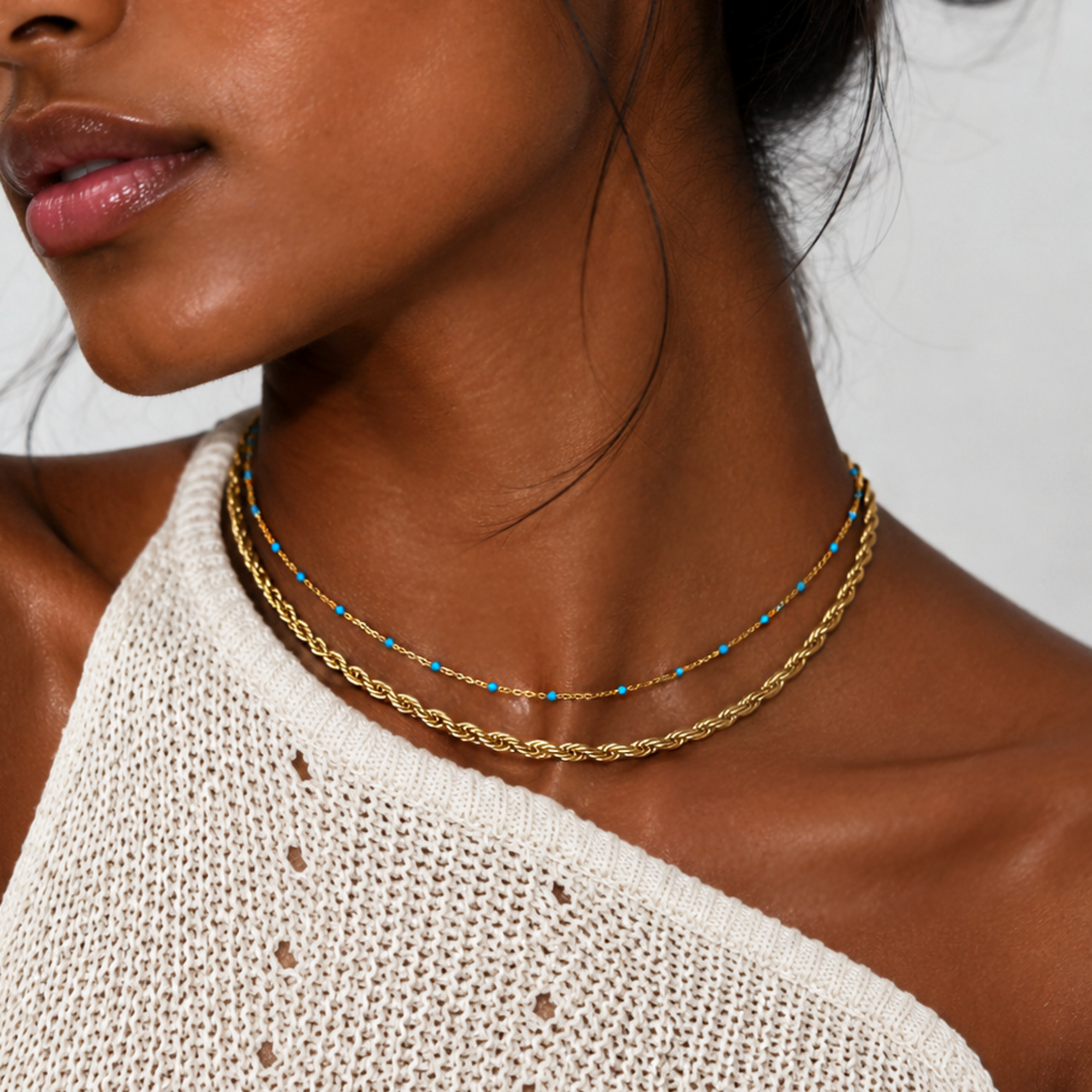 Close-up of a woman wearing a gold rope chain necklace stacked with a dainty blue beaded gold chain necklace 