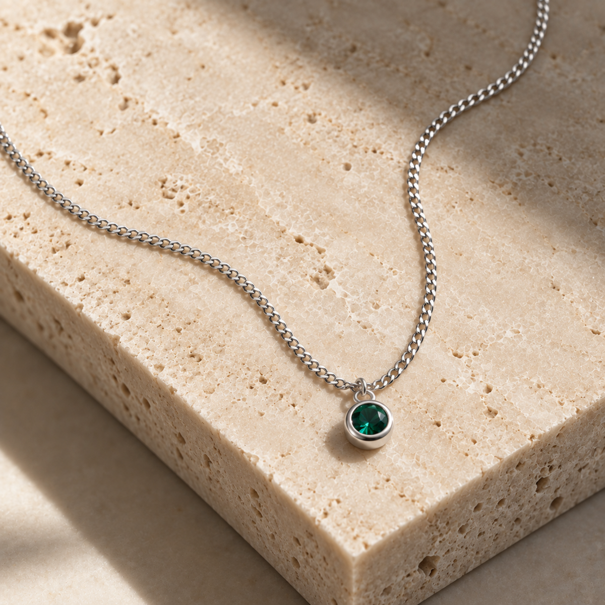 A silver chain necklace with a small round bezel-set green gemstone pendant, displayed on a textured beige stone slab.