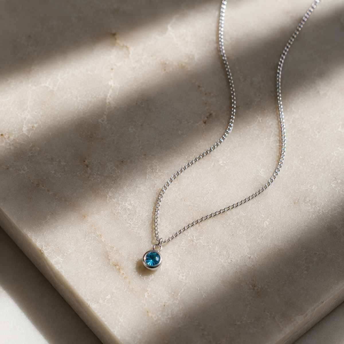 A delicate silver chain necklace with a small round blue gemstone pendant, resting on a beige stone slab with soft shadows.