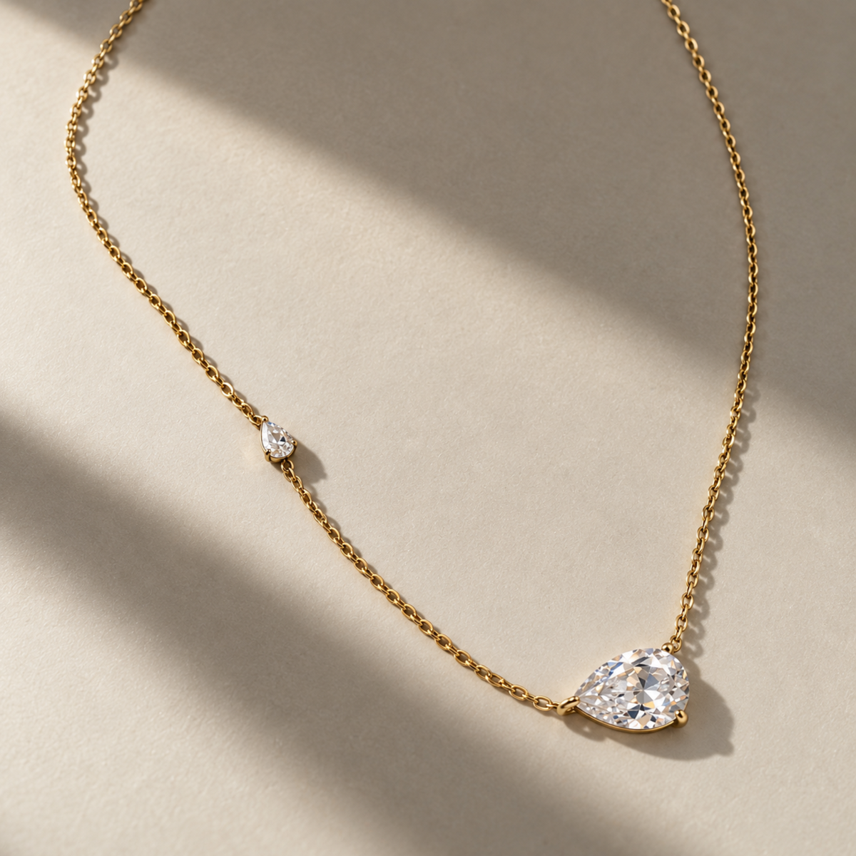 A delicate gold chain necklace featuring a pear-cut clear gemstone pendant and a smaller matching gemstone accent, displayed on a warm beige surface with soft diagonal shadows.