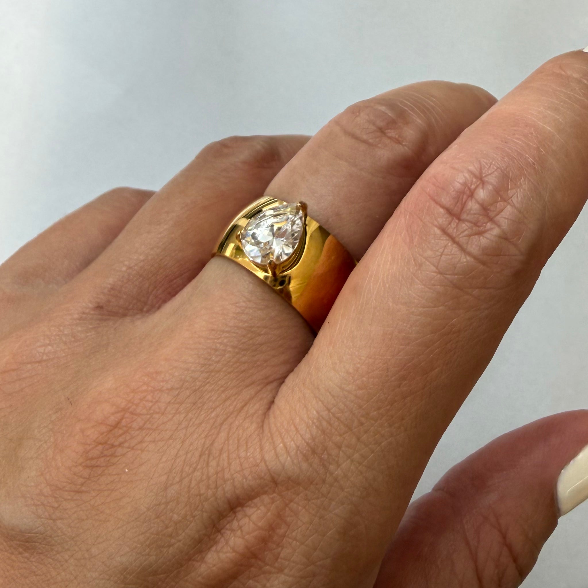 Gold ring with a pear diamond on a person's finger against a plain background