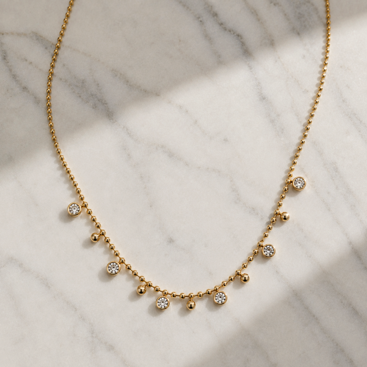 A delicate gold necklace with small bead-like links and several clear round stones set along the front, displayed on a white marble surface with soft diagonal shadows.