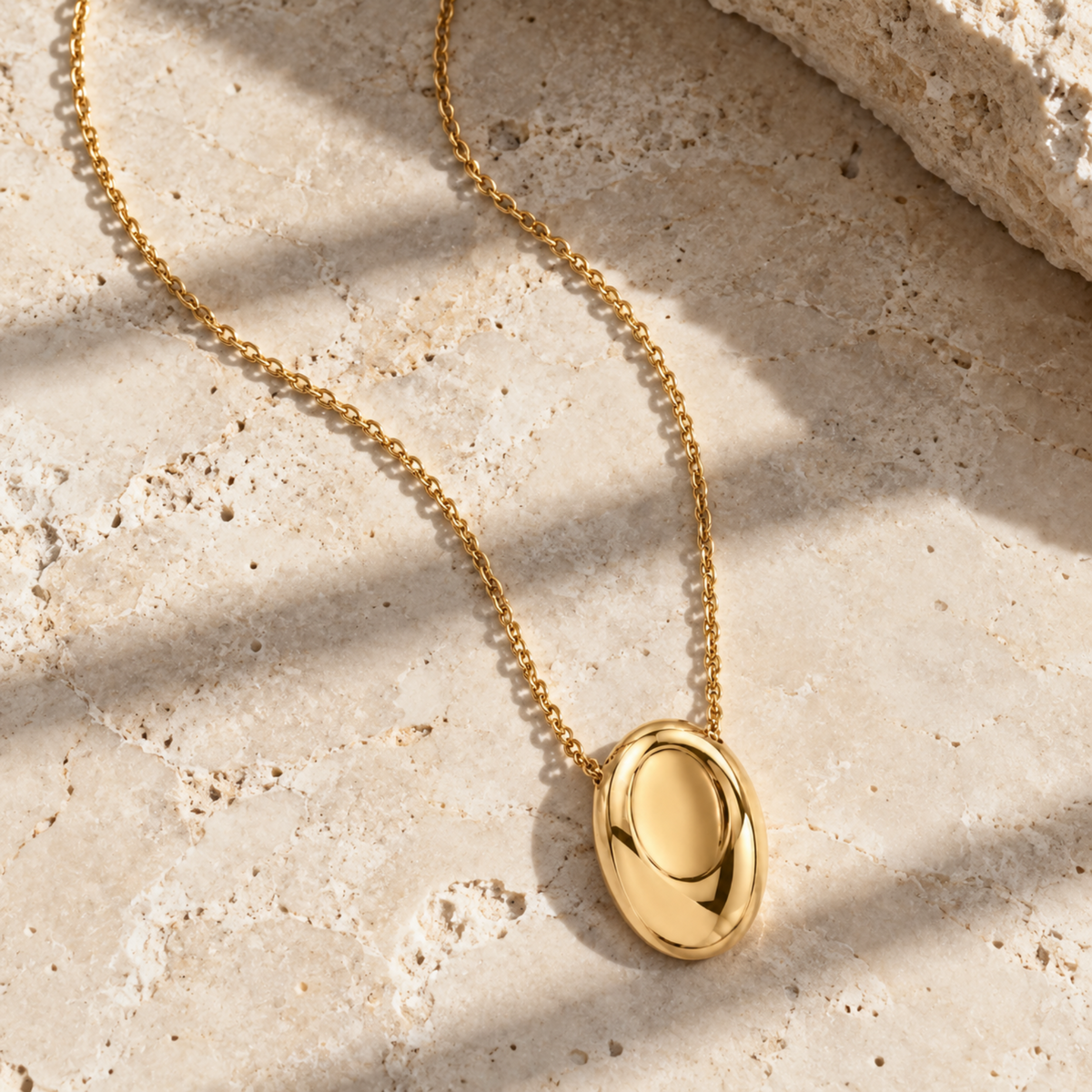 A polished gold oval pendant necklace on a delicate gold chain, displayed on a textured beige stone surface with soft diagonal shadows.