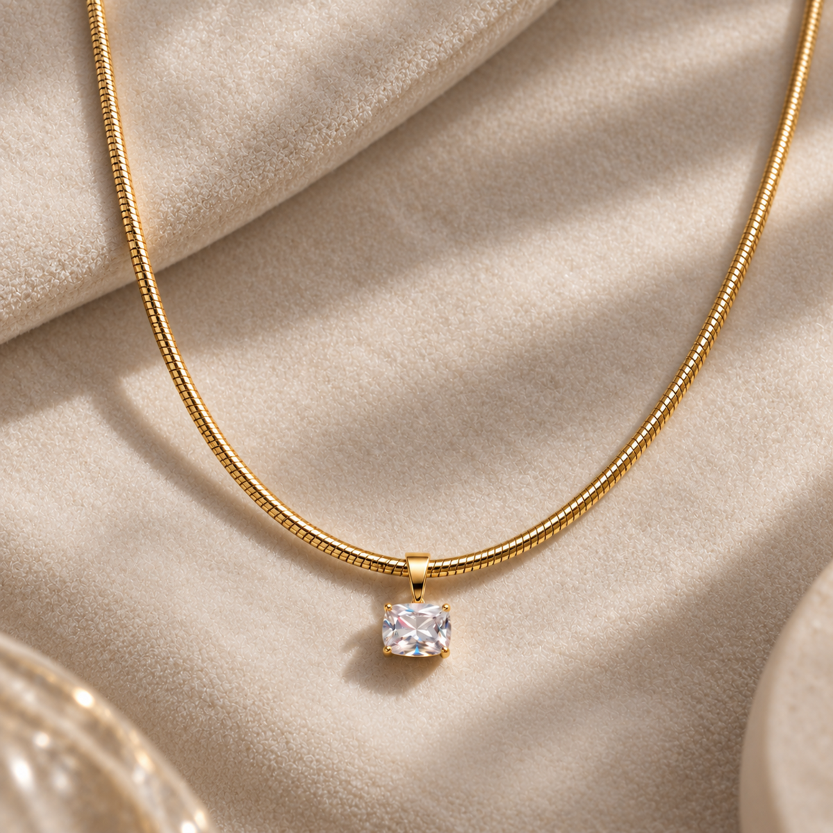 A gold snake chain necklace with a single square clear gemstone pendant, displayed on a soft beige fabric background.