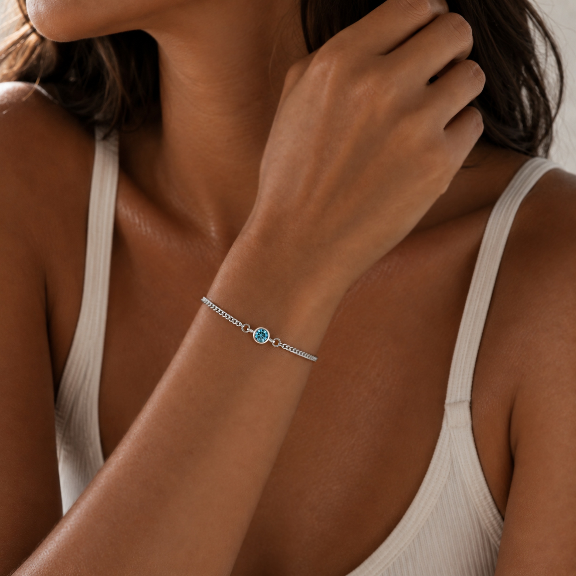 A close-up of a woman wearing a delicate silver chain bracelet with a small central blue gemstone, paired with a white ribbed tank top.