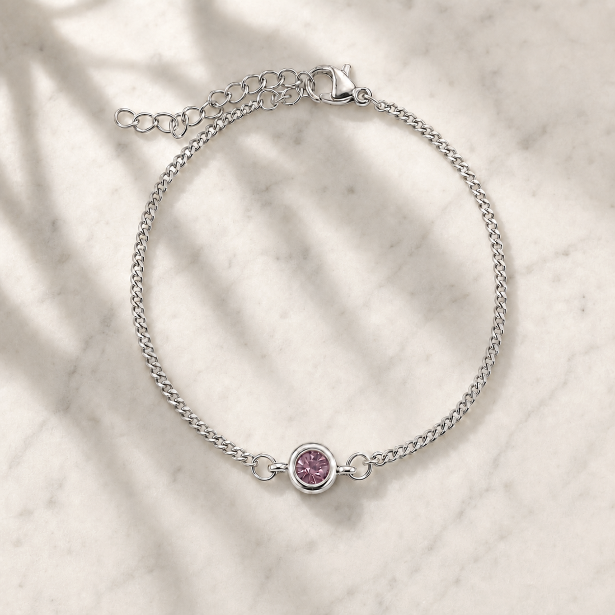 A delicate silver chain bracelet with a small round bezel-set pink gemstone, displayed on a light marble surface with soft leaf shadows.