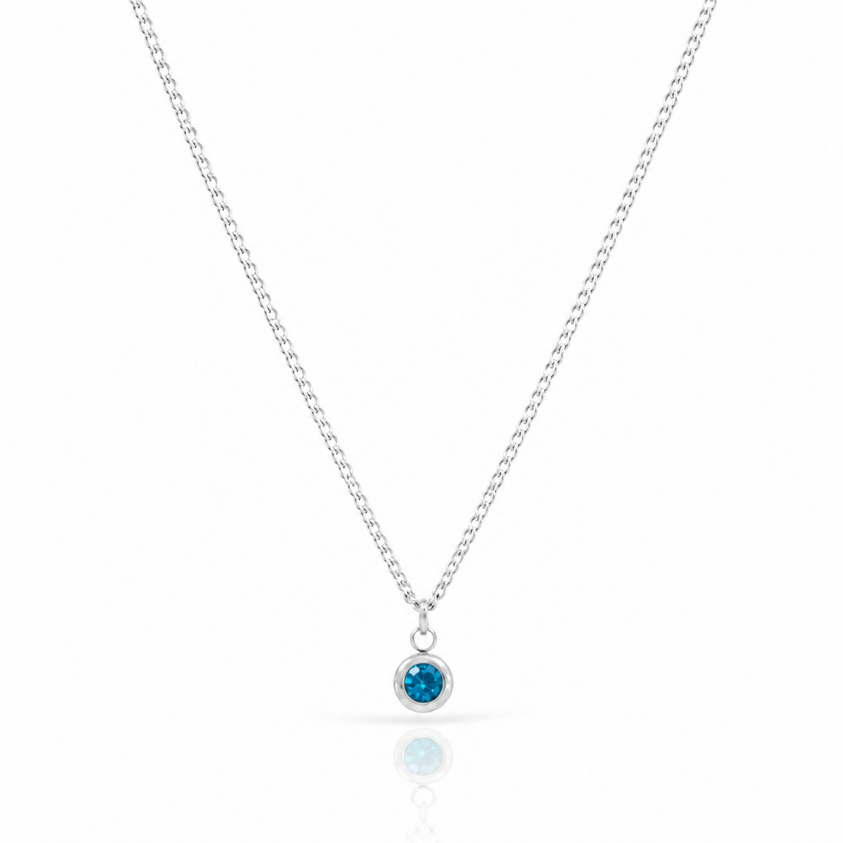 A delicate silver chain necklace with a small round bezel pendant featuring a vibrant blue gemstone, displayed on a clean white background with a soft reflection below.