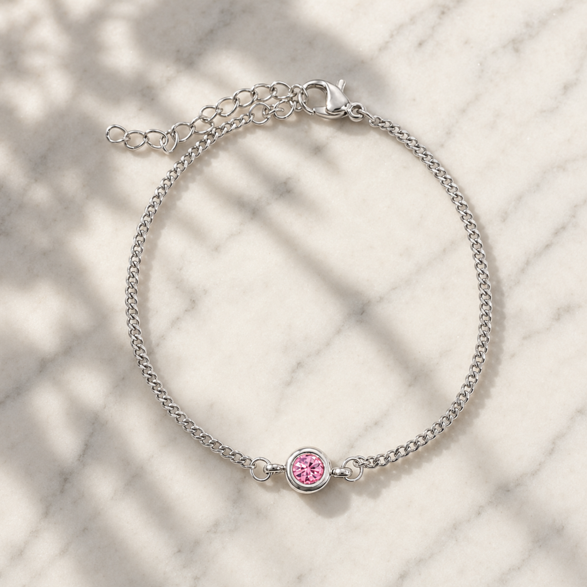 A delicate silver chain bracelet with a small round pink gemstone charm, displayed on a white marble surface with soft leaf shadows.