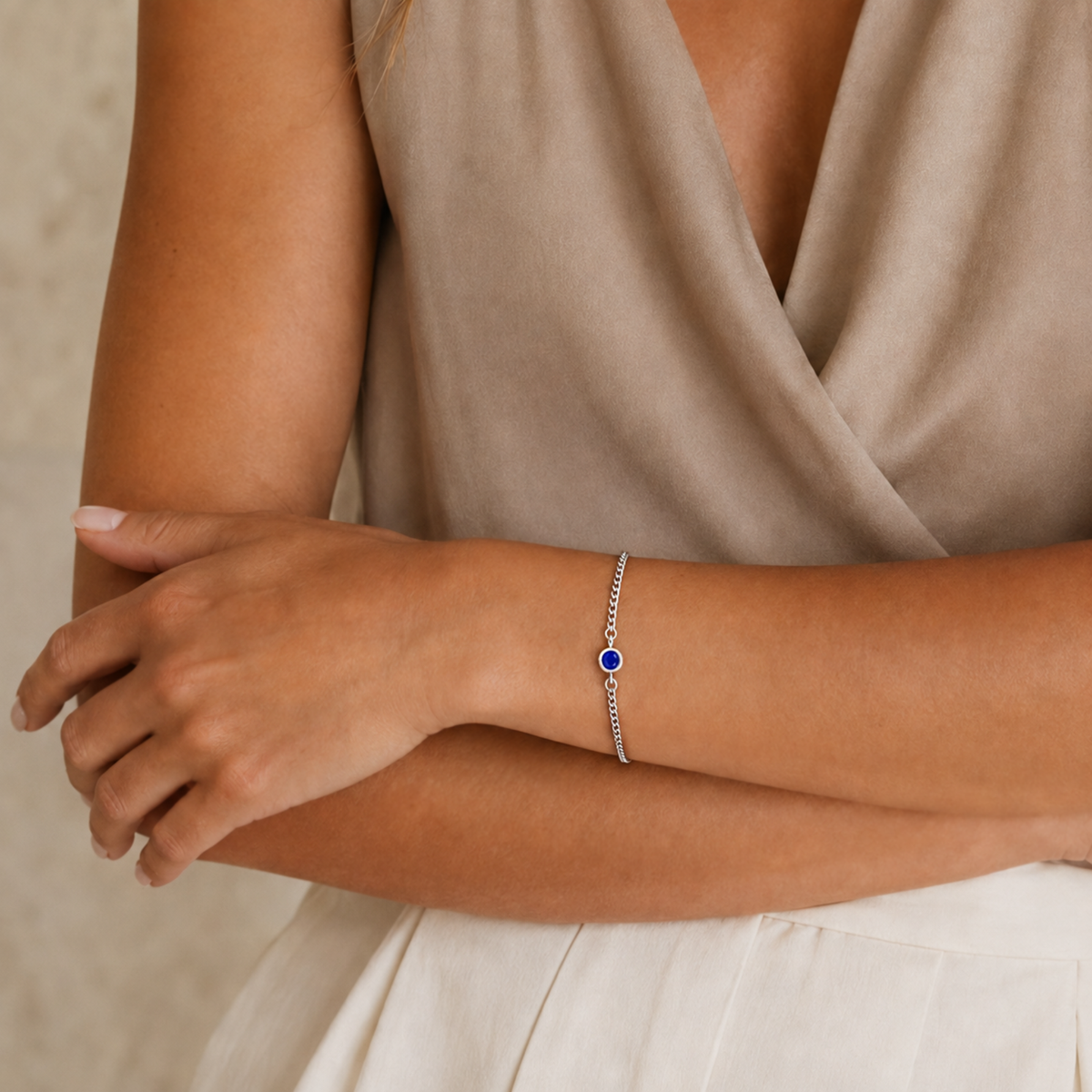 A close-up of a woman's wrist wearing a delicate silver chain bracelet with a central round blue stone, paired with a beige wrap top.