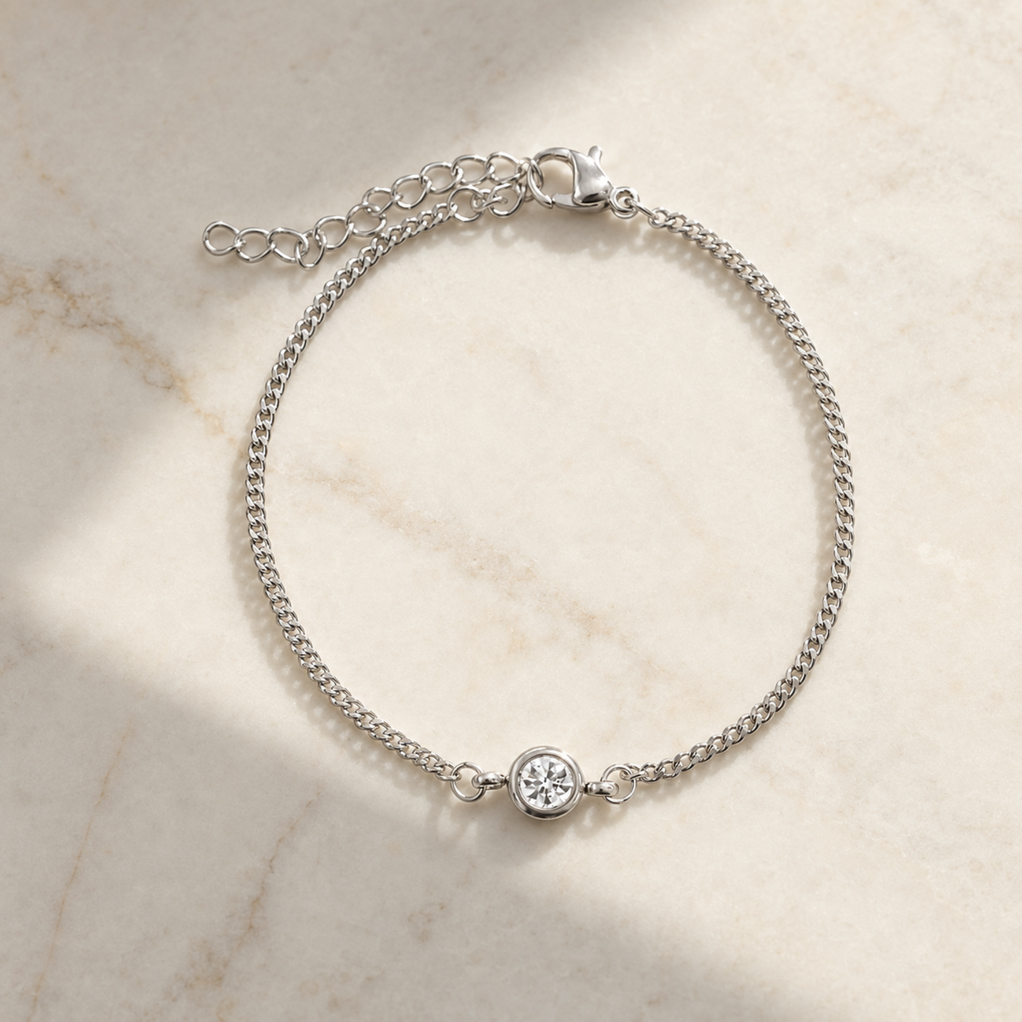A silver chain bracelet with a small round bezel-set clear stone, displayed on a light beige marble surface with soft shadows.