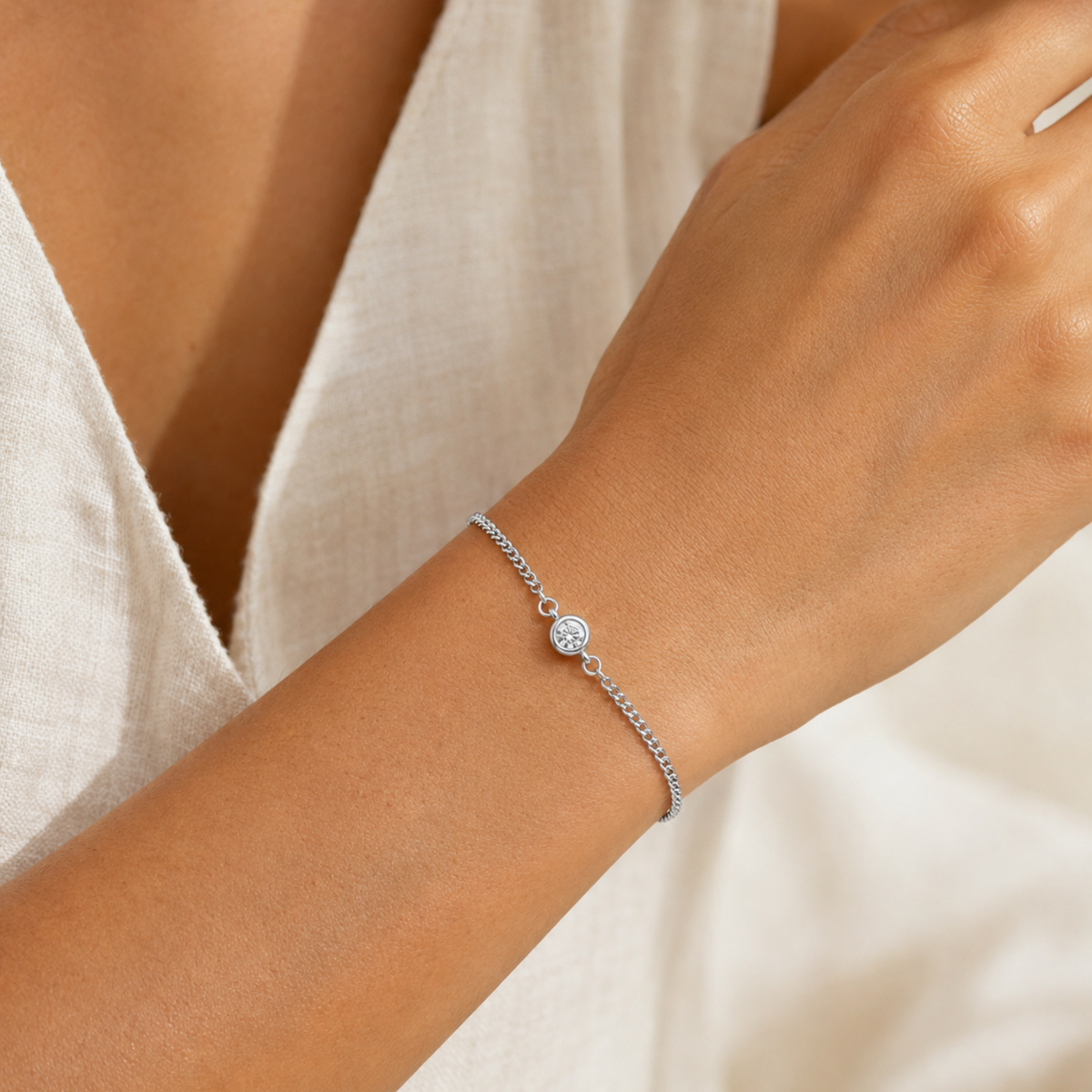 A delicate silver chain bracelet with a small round bezel-set clear stone, shown worn on a person's wrist. The person is wearing a soft, off-white linen top, and the background is a warm, softly blurred neutral tone.