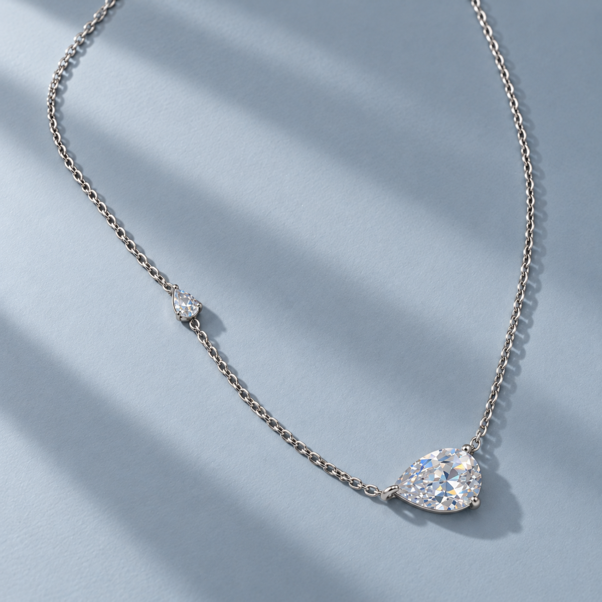 A delicate silver-tone chain necklace featuring a pear-shaped cluster of clear gemstones and a small heart-shaped stone, displayed on a light blue-gray textured background with soft diagonal shadows.