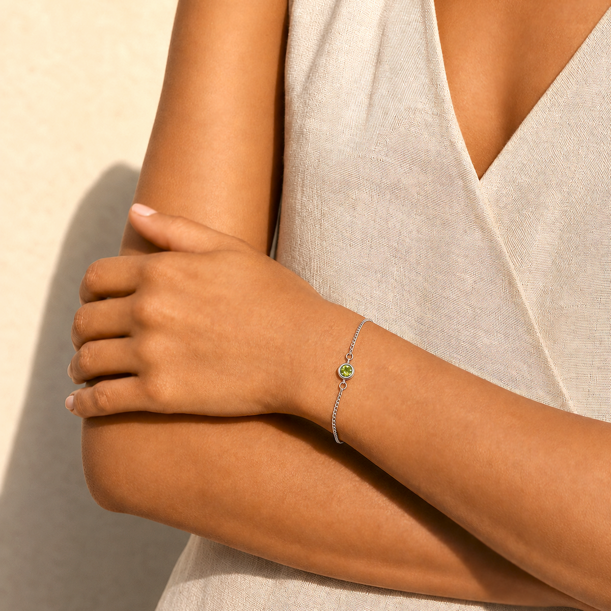 A close-up of a model's arm wearing a delicate silver chain bracelet with a small green circular charm, set against a neutral beige background.