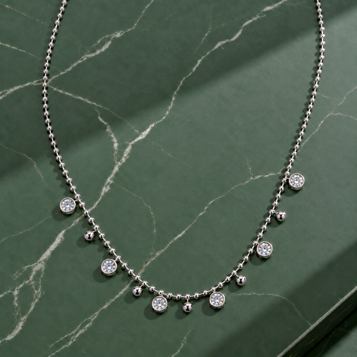 A silver ball-chain necklace with small dangling beads and several round clear stone accents, arranged on a dark green marble surface.