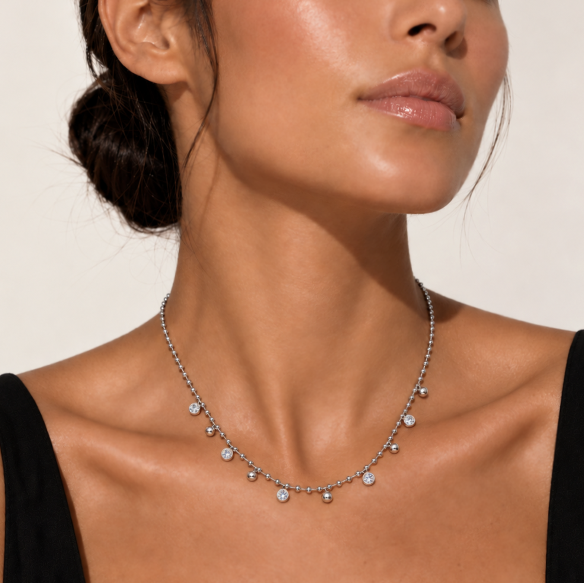 A close-up of a model wearing a delicate silver-toned beaded necklace with small clear gemstone charms, styled with a black top against a warm beige background.