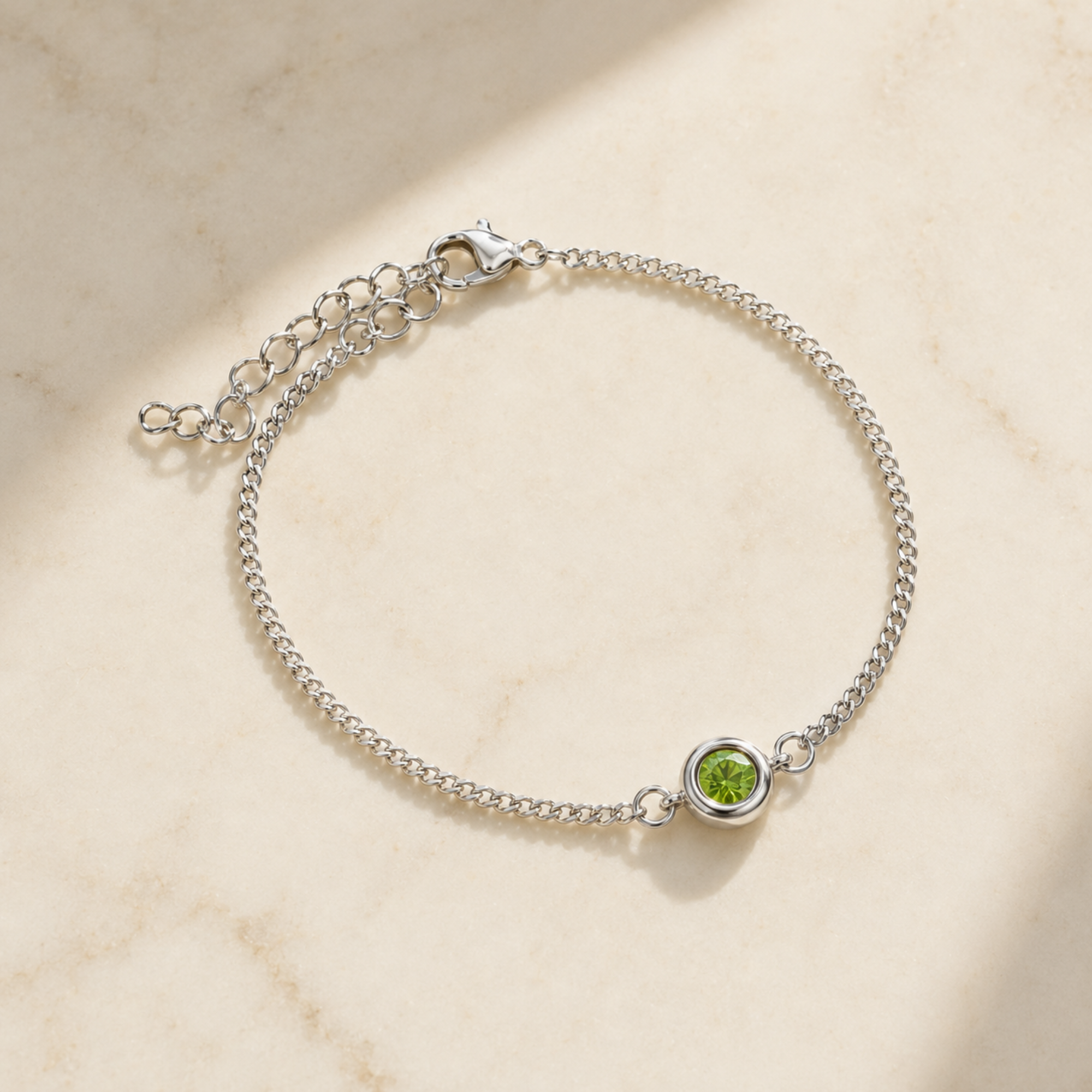 A silver chain bracelet with a small round bezel-set green gemstone, displayed on a light beige marble surface with soft shadows.