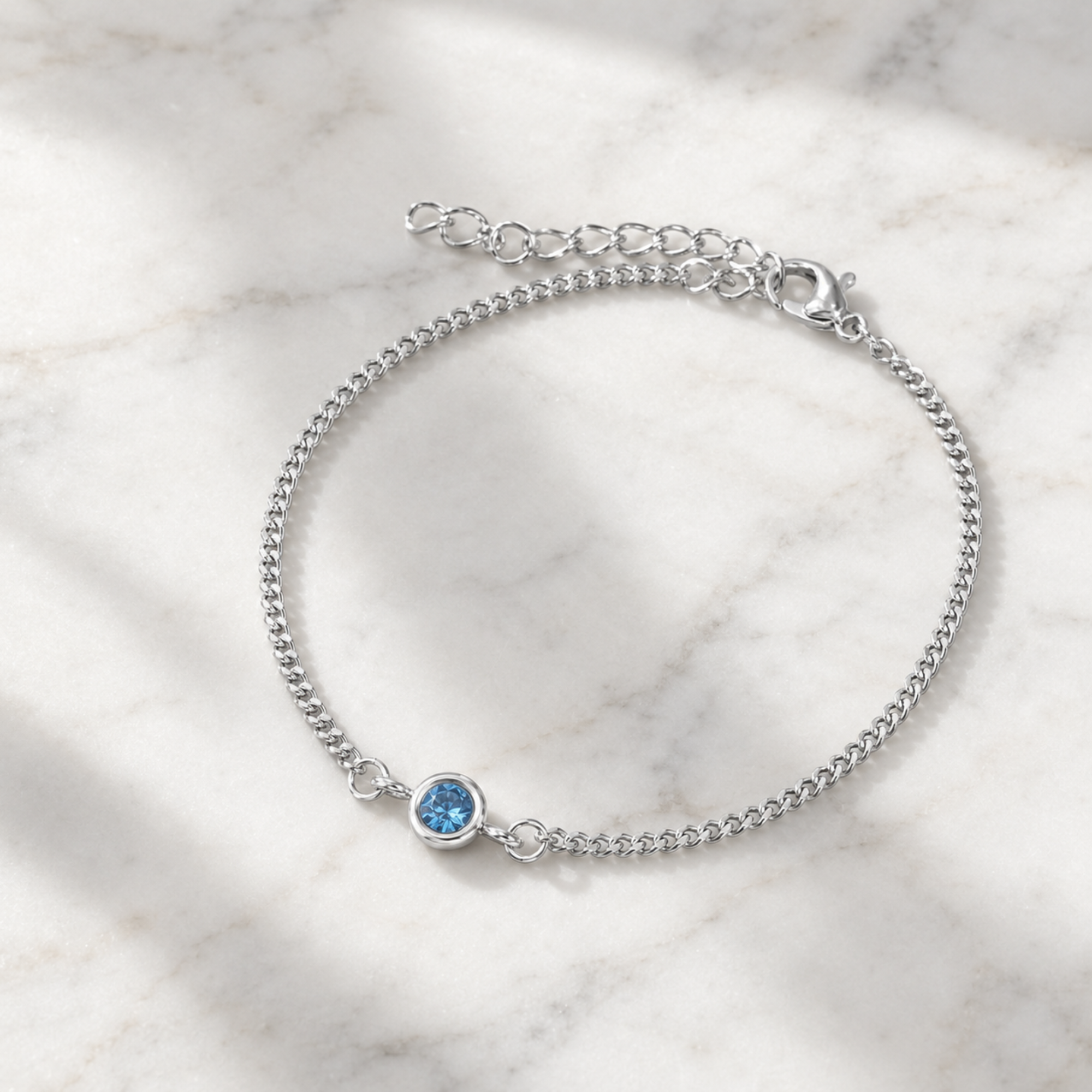 A delicate silver chain bracelet with a small round blue gemstone charm, displayed on a light marble surface with soft shadows.