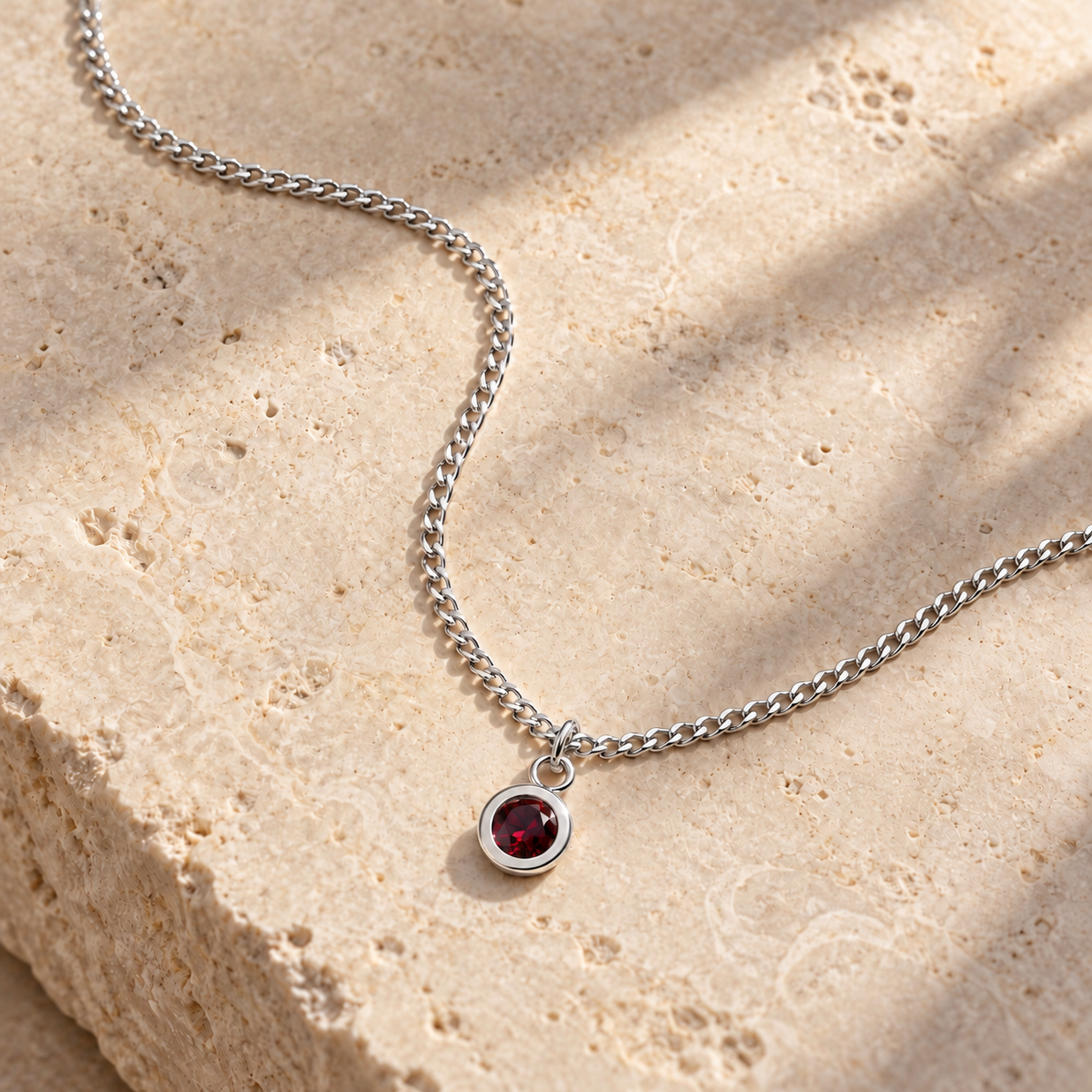 A silver chain necklace with a small round bezel-set dark red gemstone pendant, displayed on a textured beige stone surface with soft diagonal shadows.
