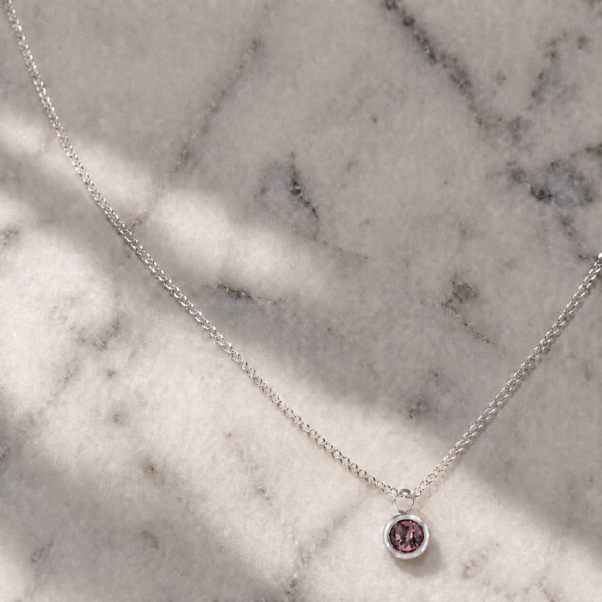 A delicate silver chain necklace with a small round bezel pendant featuring a pink gemstone, displayed on a light gray marble surface with soft shadows.