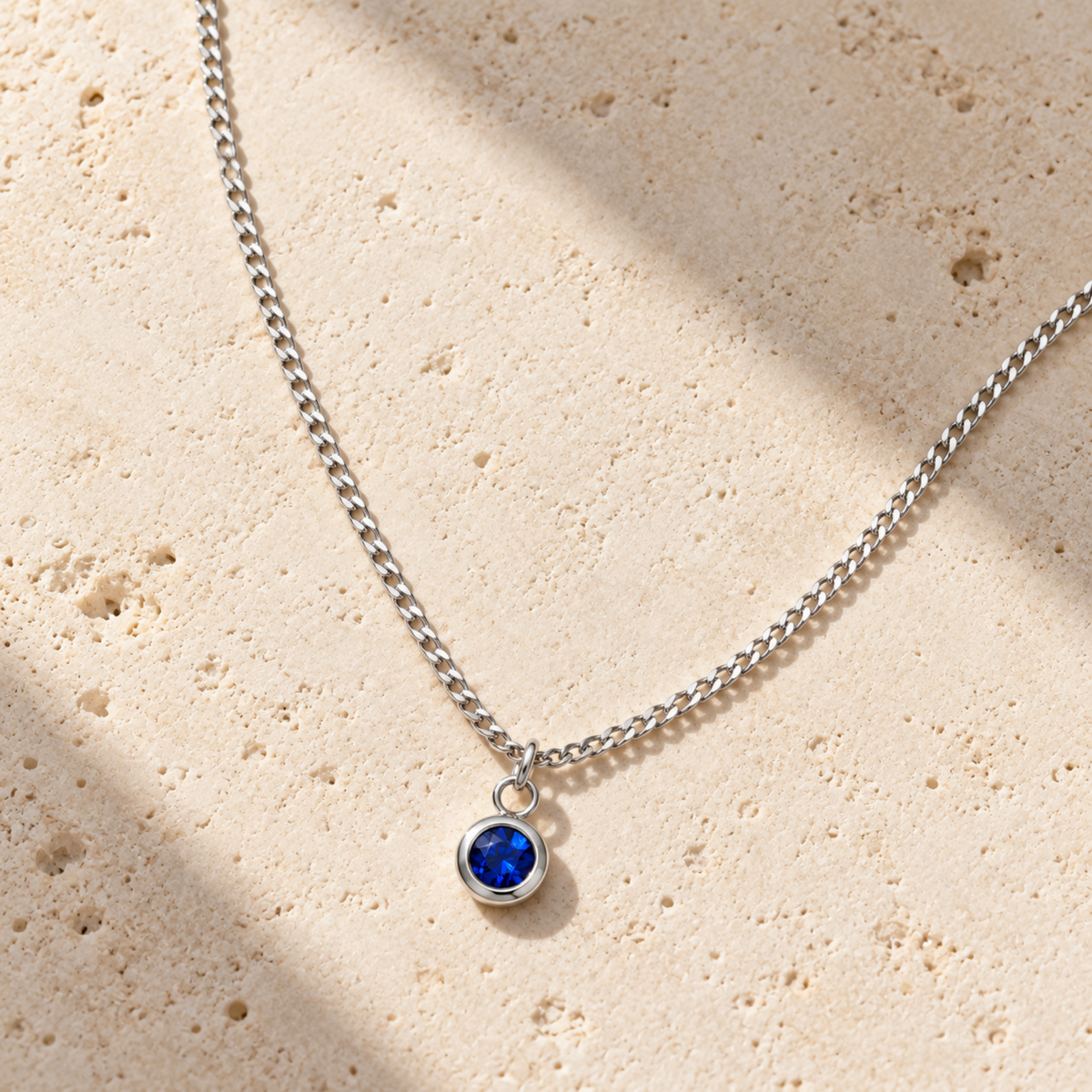 A silver chain necklace with a small round bezel-set deep blue gemstone pendant, displayed on a textured beige stone surface with soft shadows. September birthstone jewelry 