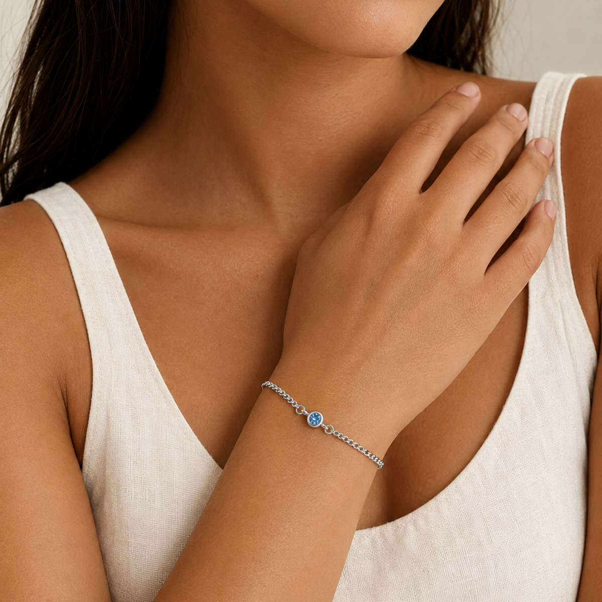 A close-up of a model wearing a delicate silver chain december birthstone bracelet with a central round blue gemstone, paired with a white tank top.