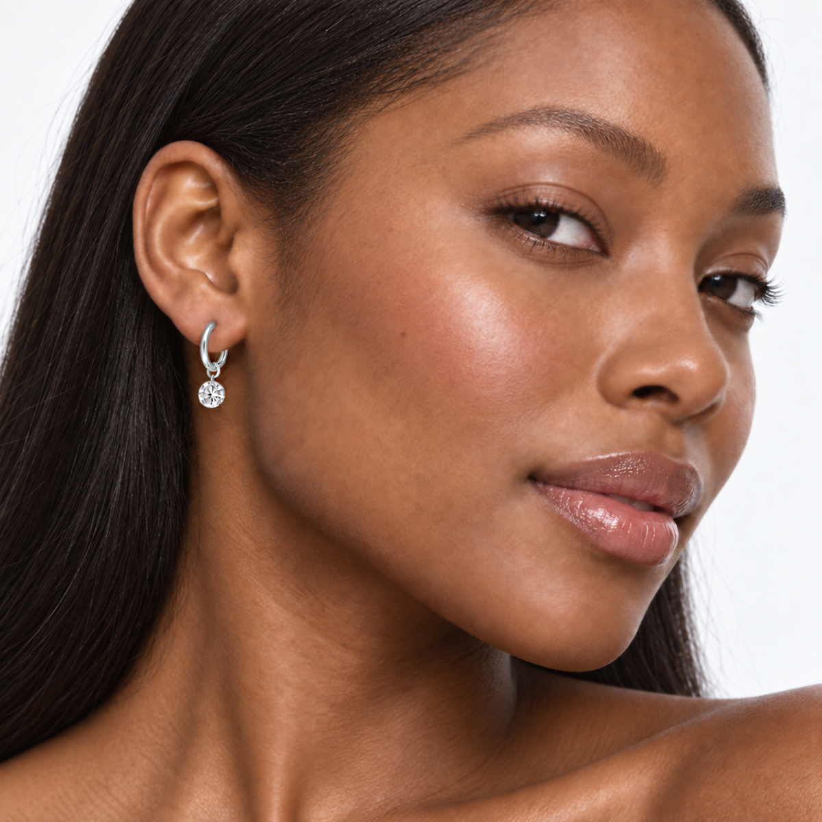 A close-up of a woman wearing a small silver hoop earring with a clear round gemstone dangle, shown against a clean white background.
