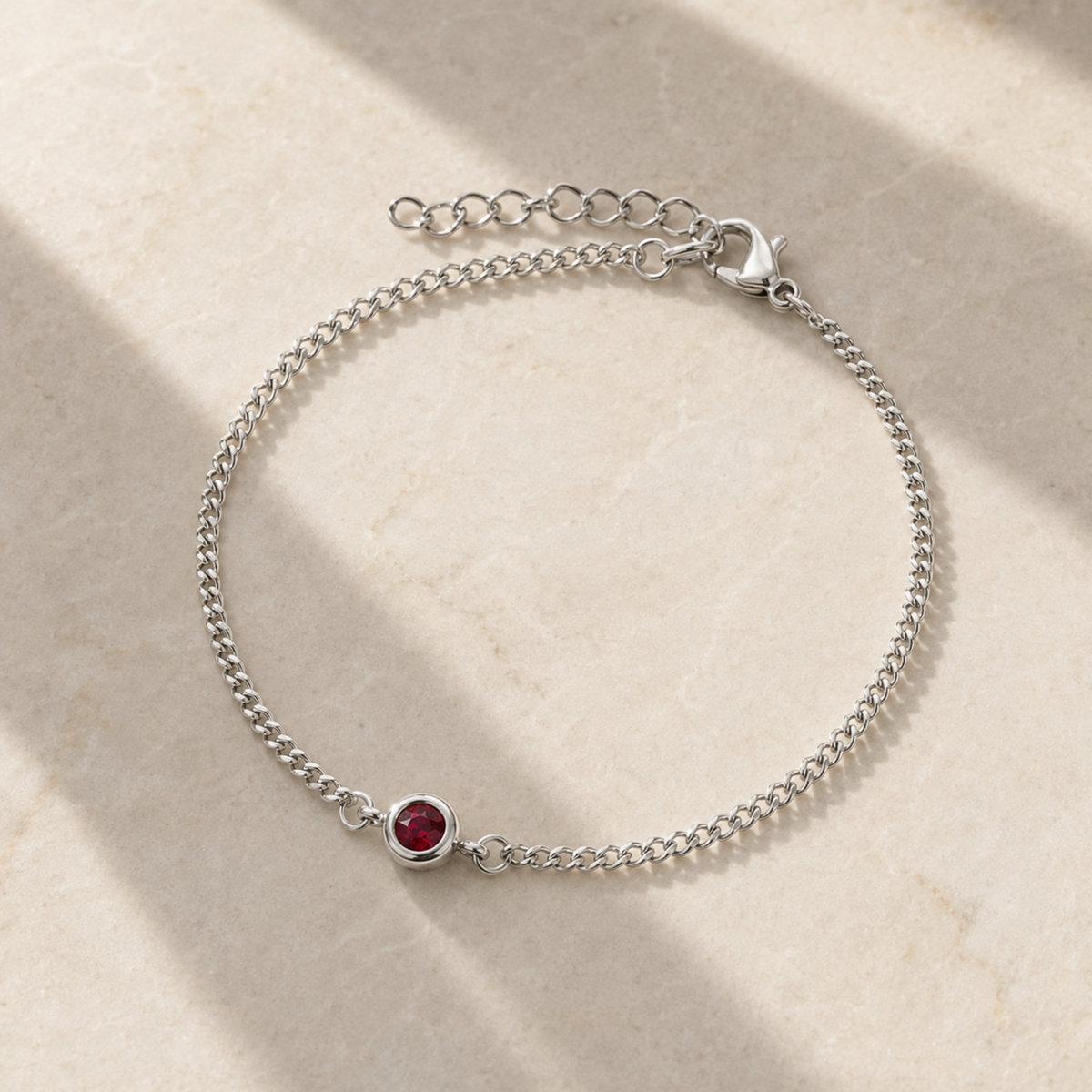 A delicate silver chain bracelet with a single bezel-set round red gemstone, displayed on a light beige textured surface with soft diagonal shadows.