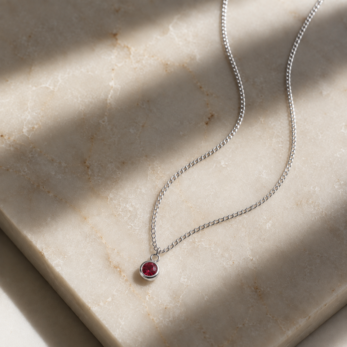 A delicate silver chain necklace with a small round blue gemstone pendant, resting on a beige stone slab with soft shadows.