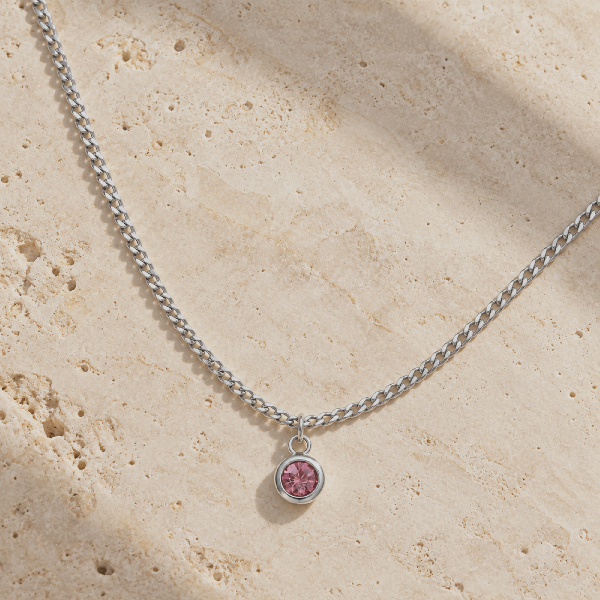 A silver-toned chain necklace with a small round bezel pendant containing a pink gemstone, displayed on a textured beige stone surface with soft shadows.