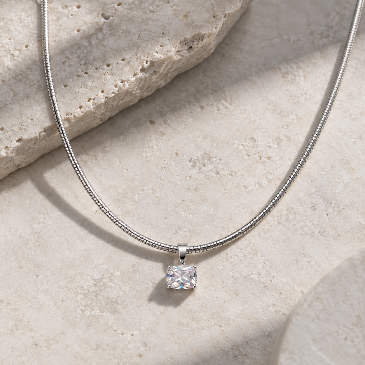 A silver snake chain necklace with a single princess-cut clear gemstone pendant, displayed on a textured beige stone surface.