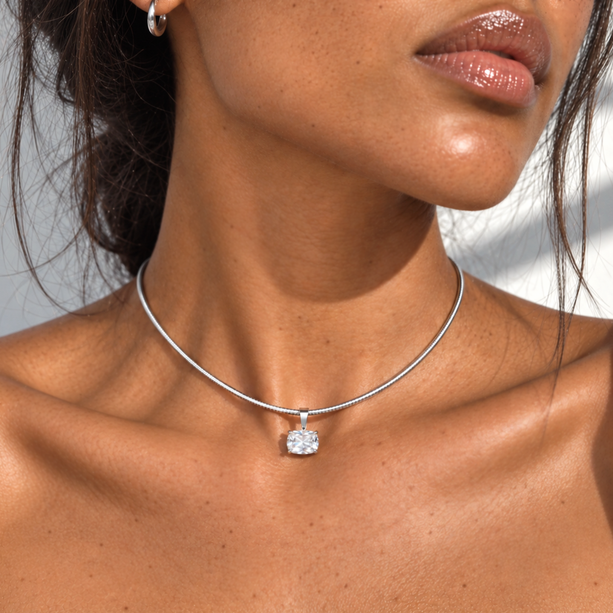 A close-up of a model's neck and chest wearing a delicate silver snake chain necklace with a single bezel-set clear gemstone pendant.