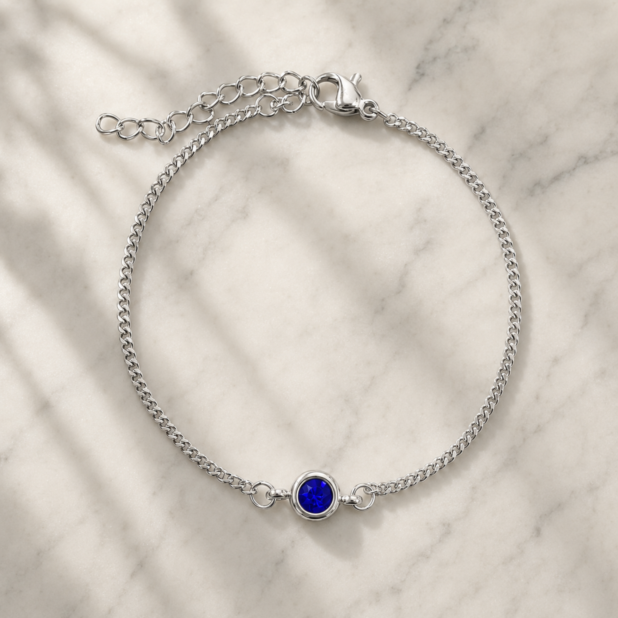 A silver chain bracelet with a small round blue gemstone centerpiece, displayed on a light marble surface with soft leaf shadows.