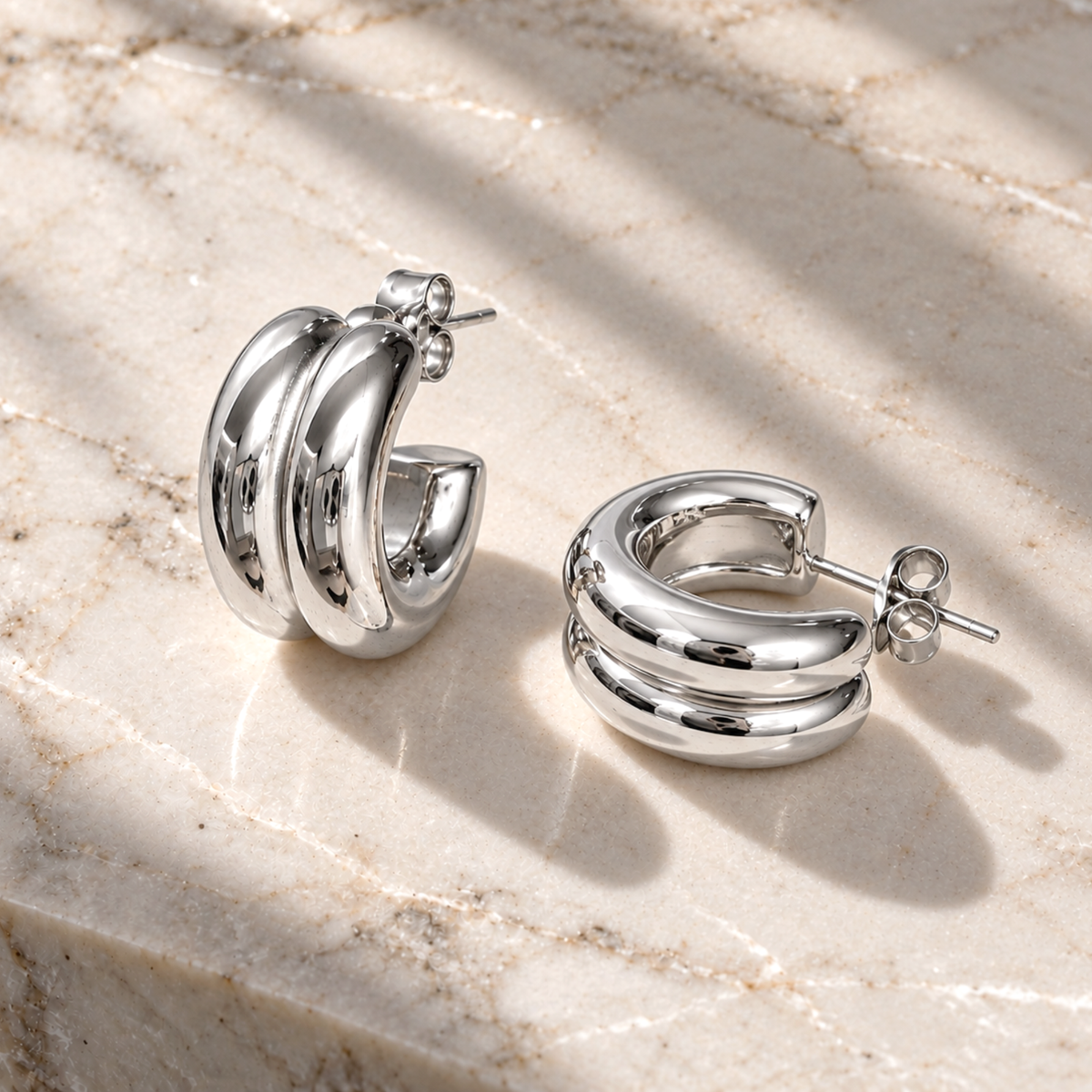 A pair of polished silver hoop earrings with three parallel curved bands, displayed on a light beige marble surface with soft shadows.