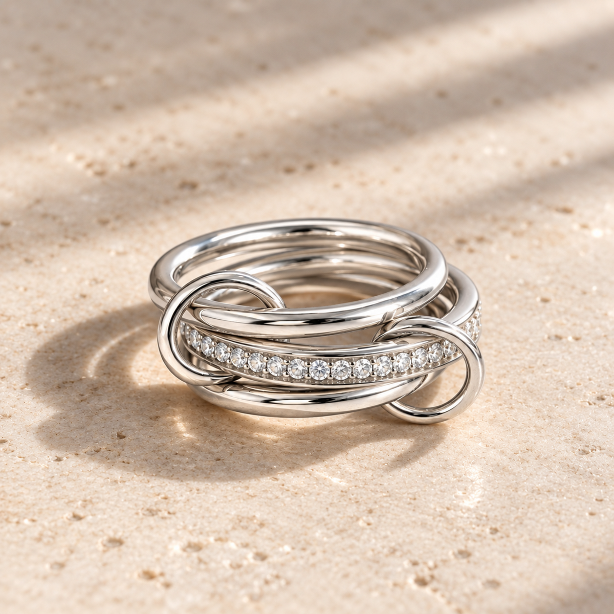 A polished silver-toned ring with three interlocking bands, one of which is paved with small clear stones, resting on a textured beige stone surface with soft shadows.
