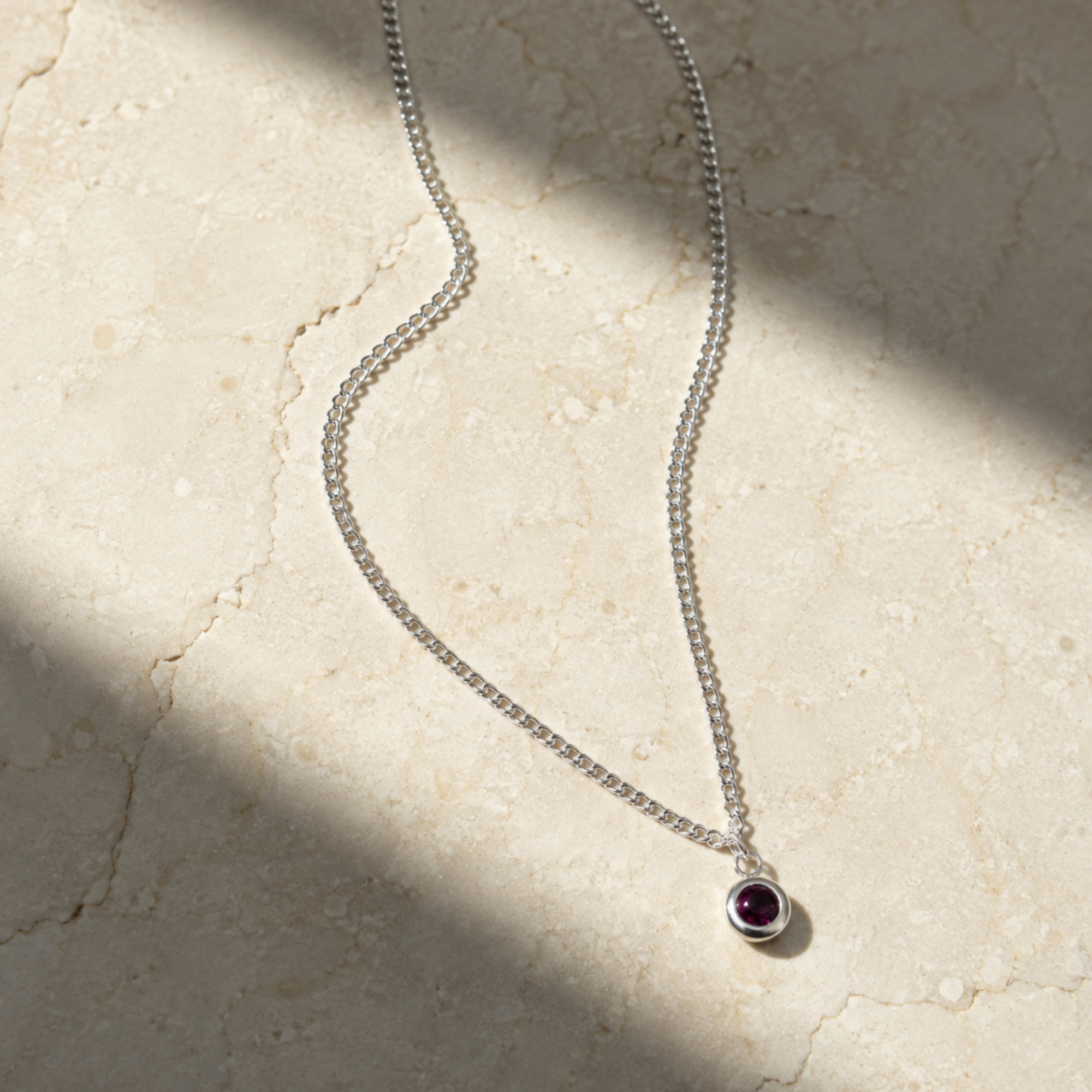 A delicate silver chain necklace with a small bezel-set round purple gemstone pendant, displayed on a textured beige stone surface with soft diagonal shadows.
