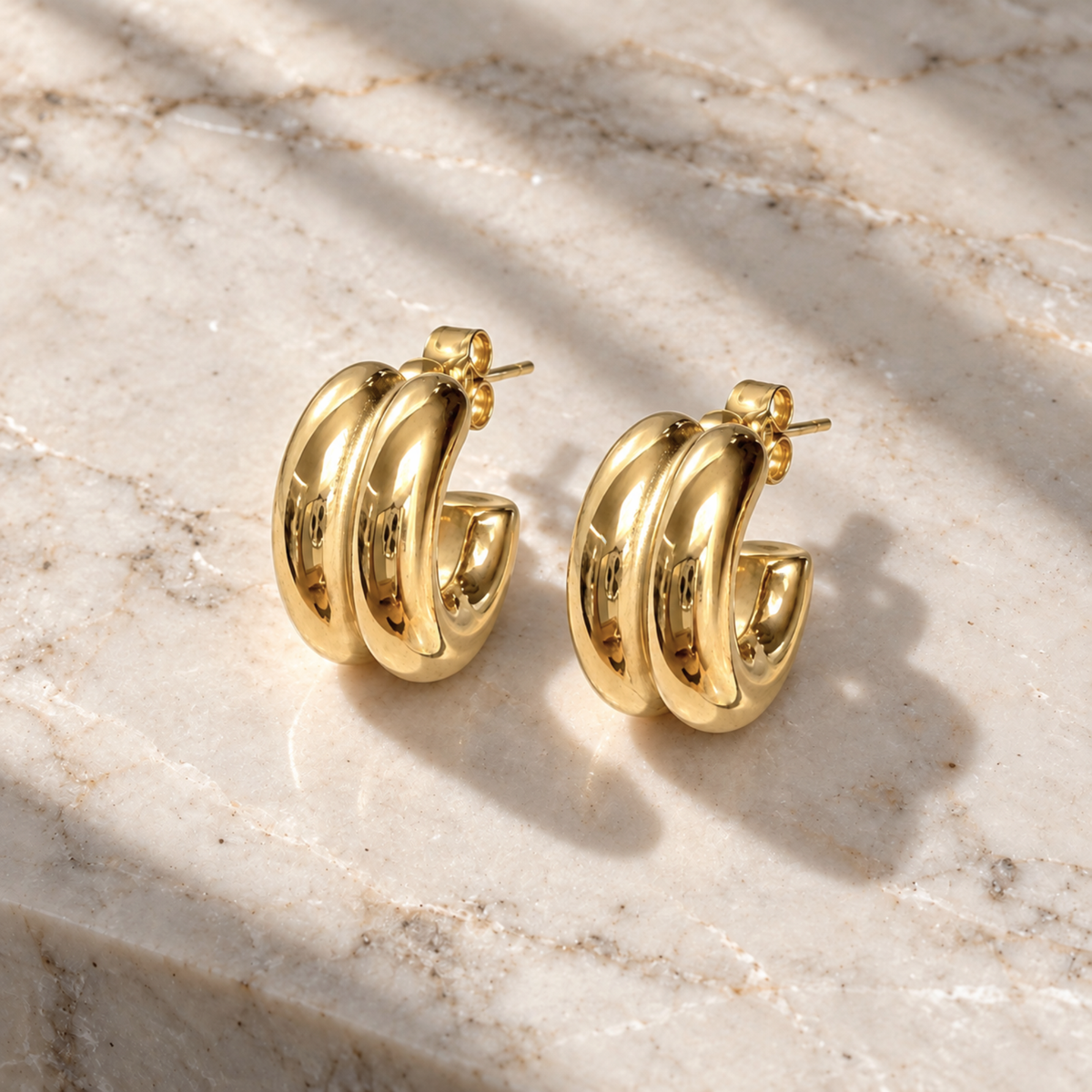 A pair of polished gold hoop earrings with a triple-loop twisted design, displayed on a light beige marble surface with soft shadows.