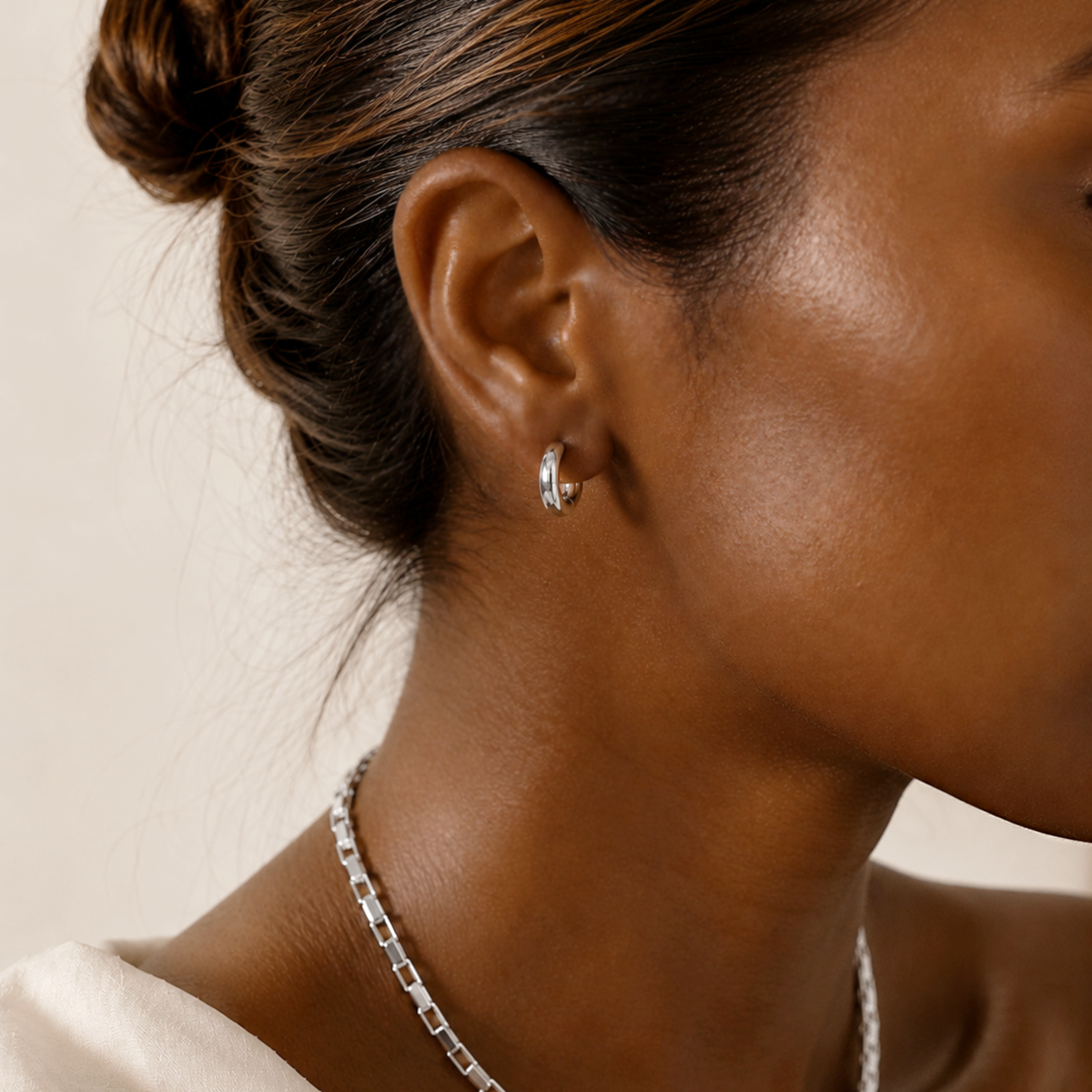 A close-up of a woman wearing a small silver hoop earring and a silver chain necklace against a soft beige background.